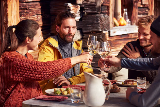 4 persone, una donna e due uomini, si siedono in una capanna e brindano con vino bianco