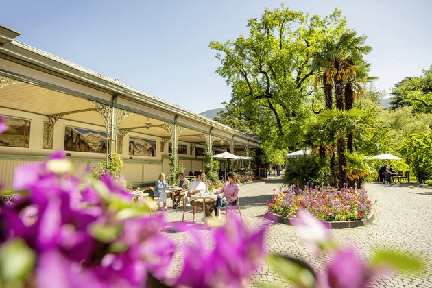 Vista sulla Wandelhalle di Merano, a fianco delle persone sedute ai tavoli si gustano un caffé in una giornata di sole, tutt'intorno fiori e una vegetazione mediterranea