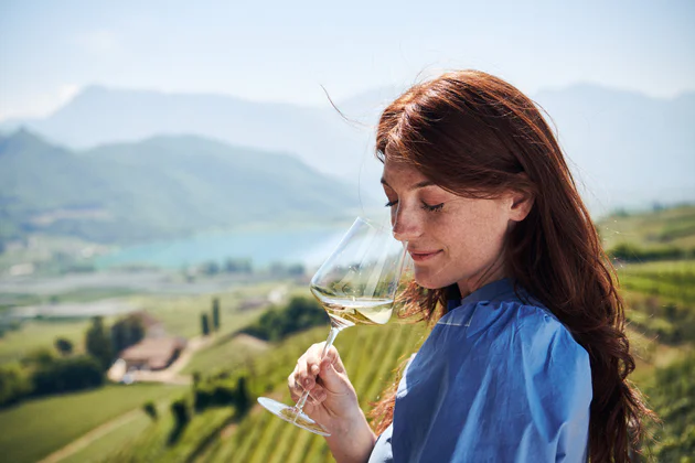Una donna si gusta un bicchiere di vino bianco con alle spalle una vista sulla Strada del Vino dell'Alto Adige, circondata da montagne, alberi e vigneti verdi.