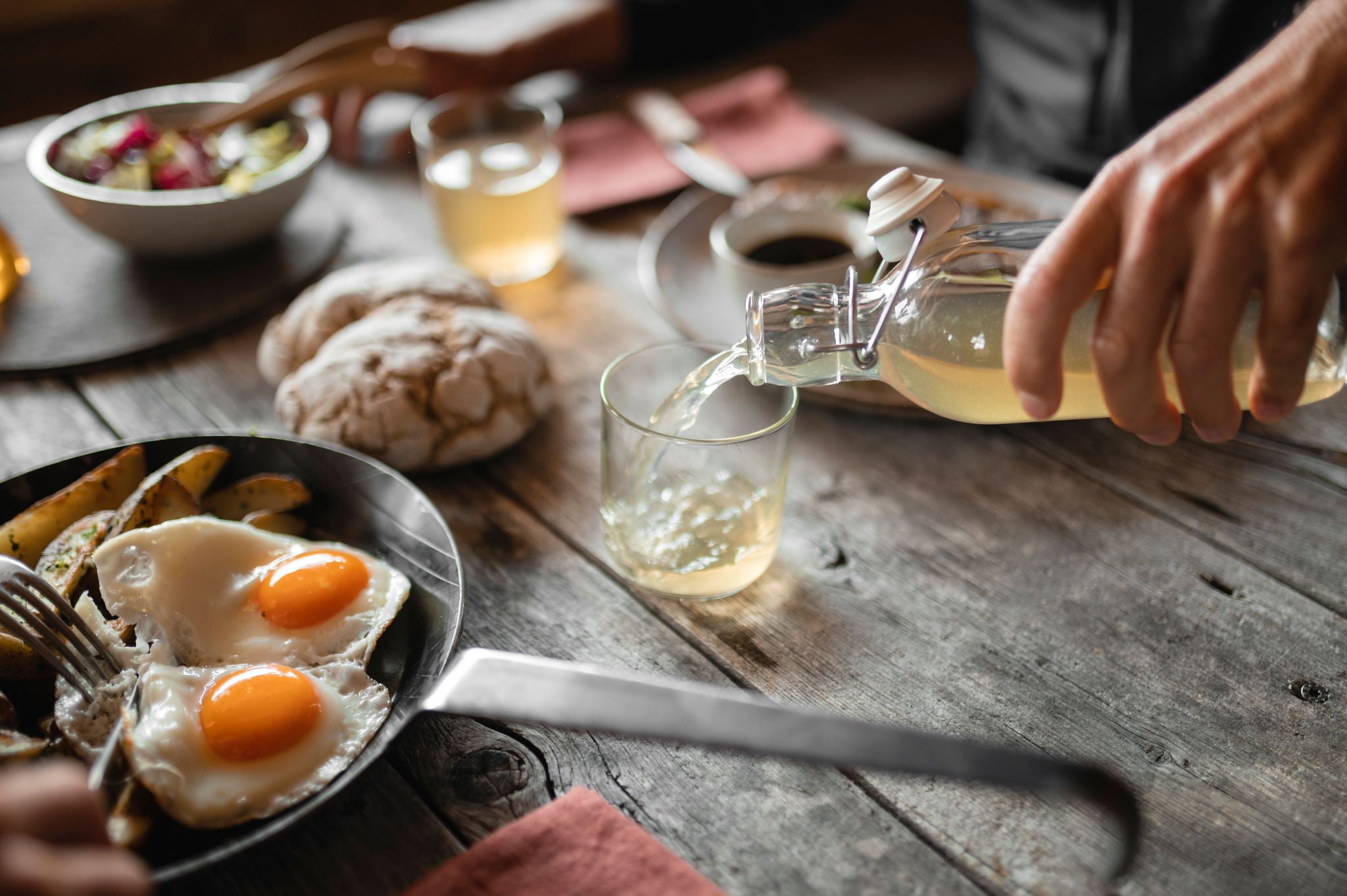Ein rustikaler Holztisch gedeckt mit Südtiroler Qualitätsprodukten, darunter Brot, Kartoffeln und Spiegeleier. Aus einer Glasflasche wird Apfelsaft in ein Glas eingeschänkt. 