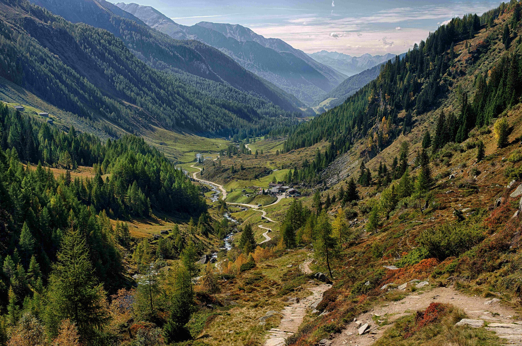 Ein kleiner Wanderweg schlängelt sich durch die bewaldeten Berghänge talwärts, wo ein kleiner Fluss und urige Almhütten die Landschaft prägen.