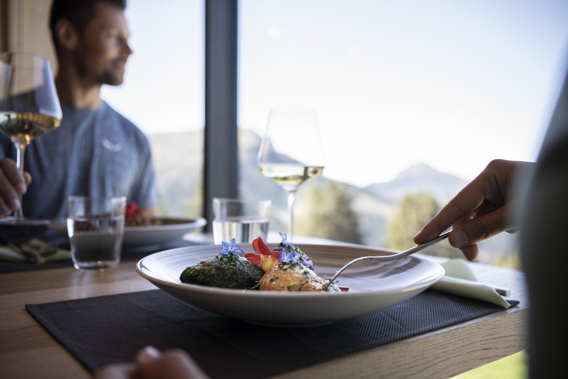 Ein Teller mit Knödel und ein Glas Wein stehen auf dem Tisch an der Fensterfront, ein Mann erhebt das Weinglas
