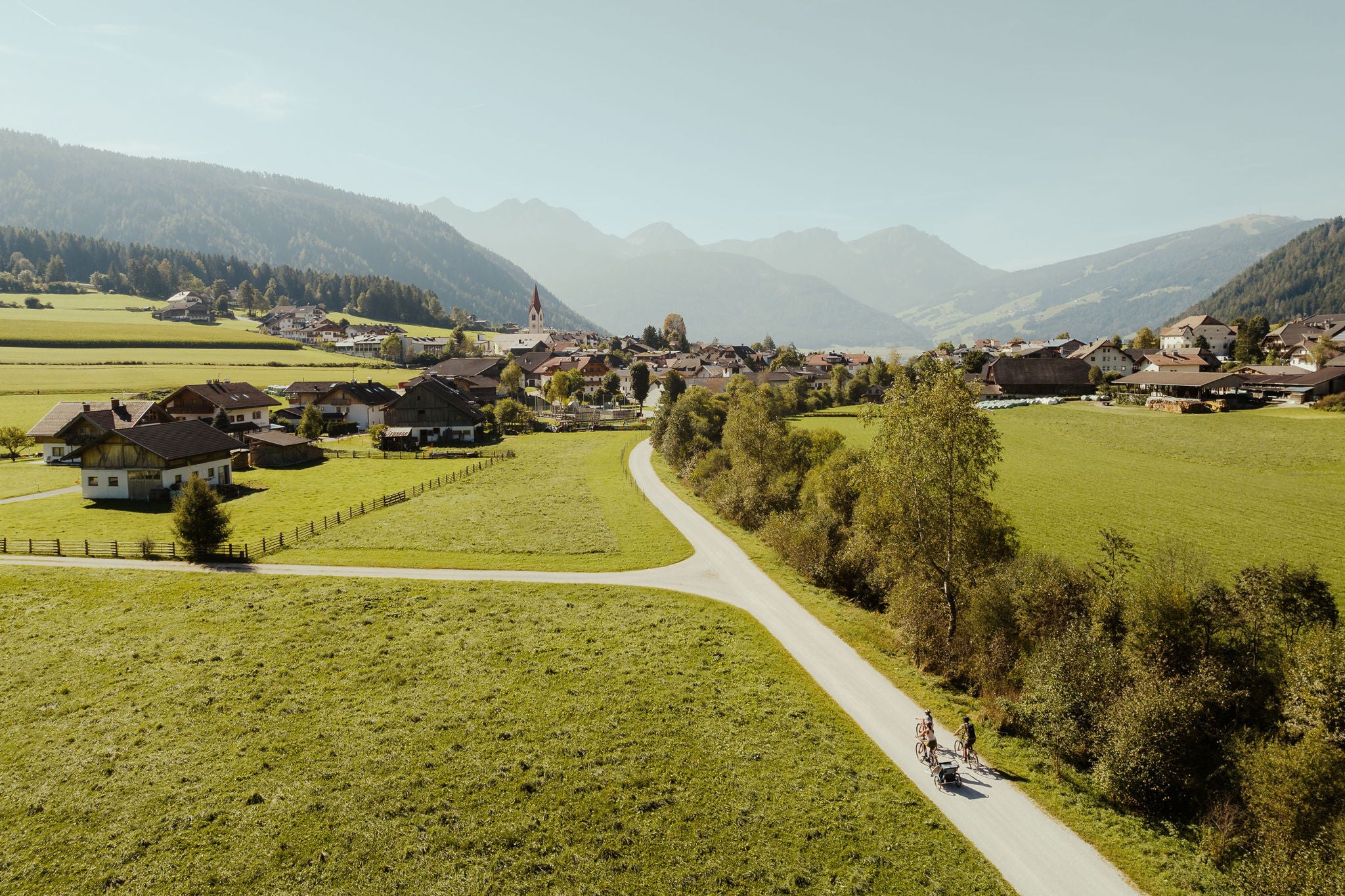 Idyllisches kleines Dorf mit Bergen und Wiesen, welche von eine Radweg durchkreuzt werden, aus der Vogelperspektive