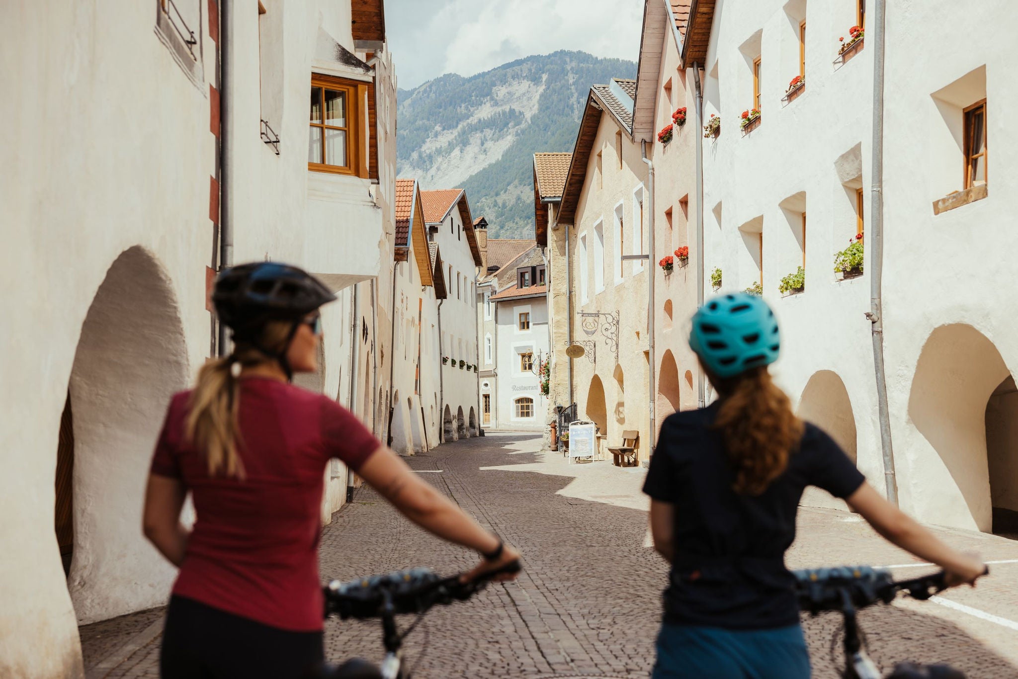 Zwei junge Radfahrerinnen schieben ihre Räder durch die gepflasterte Laubengasse von Glurns