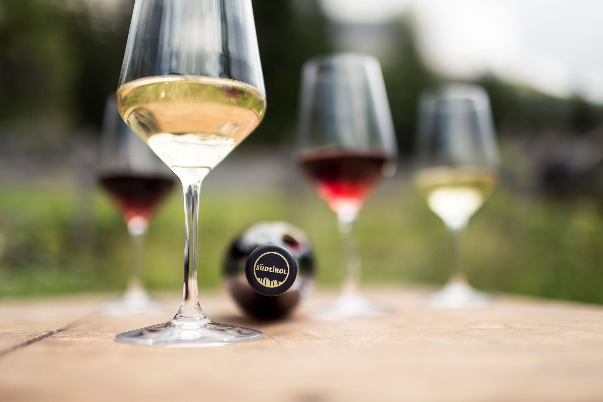 Auf einem hölzernen Untergrund stehen 4 Weingläser, ein Weinglas mit Weißwein im Vordergrund, ein weiteres und zwei Rotweingläser unscharf im Hintergrund. Zwischen den Gläsern liegt eine Weinflasche mit der Südtirol Kapsel.