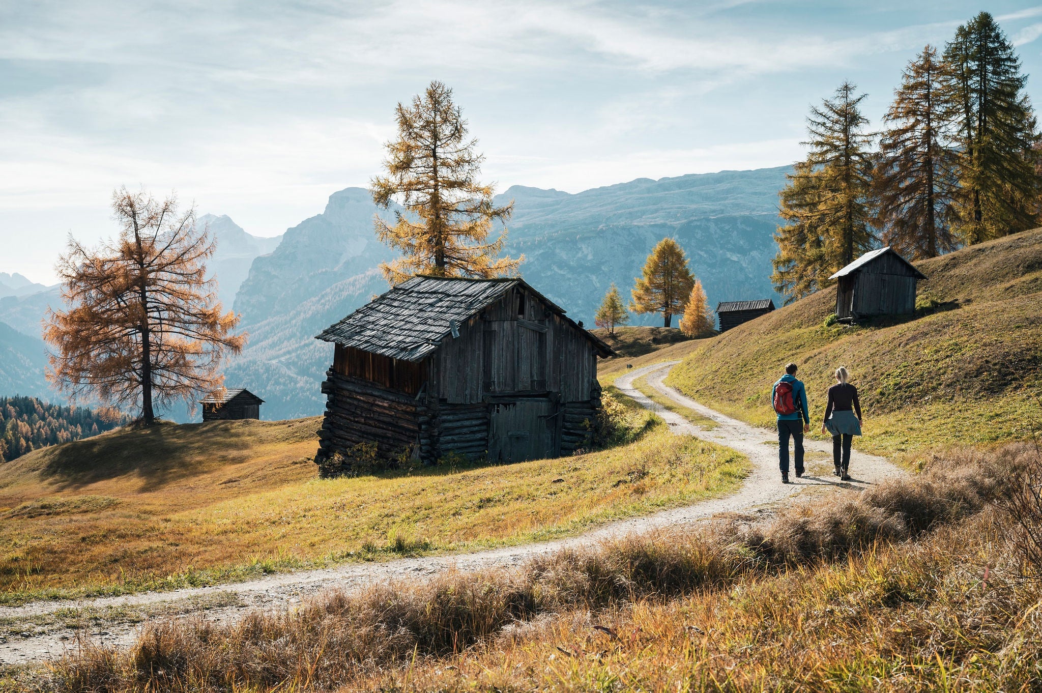 Ein Mann und eine Frau wandern an einem sonnigen Herbsttag zwischen Holzhütten über einen Almweg in der Region Alta Badia.