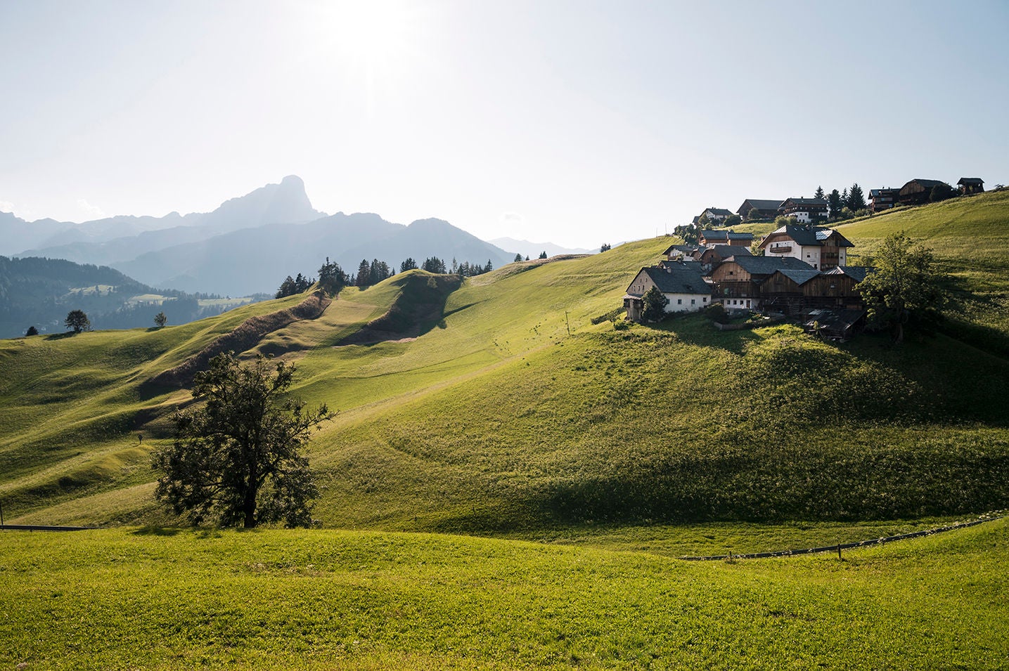 Eine Gruppe von Bauernhöfen auf einem sonnigen Berghang, dahinter erhebt sich der Berggipfel Peitlerkofel