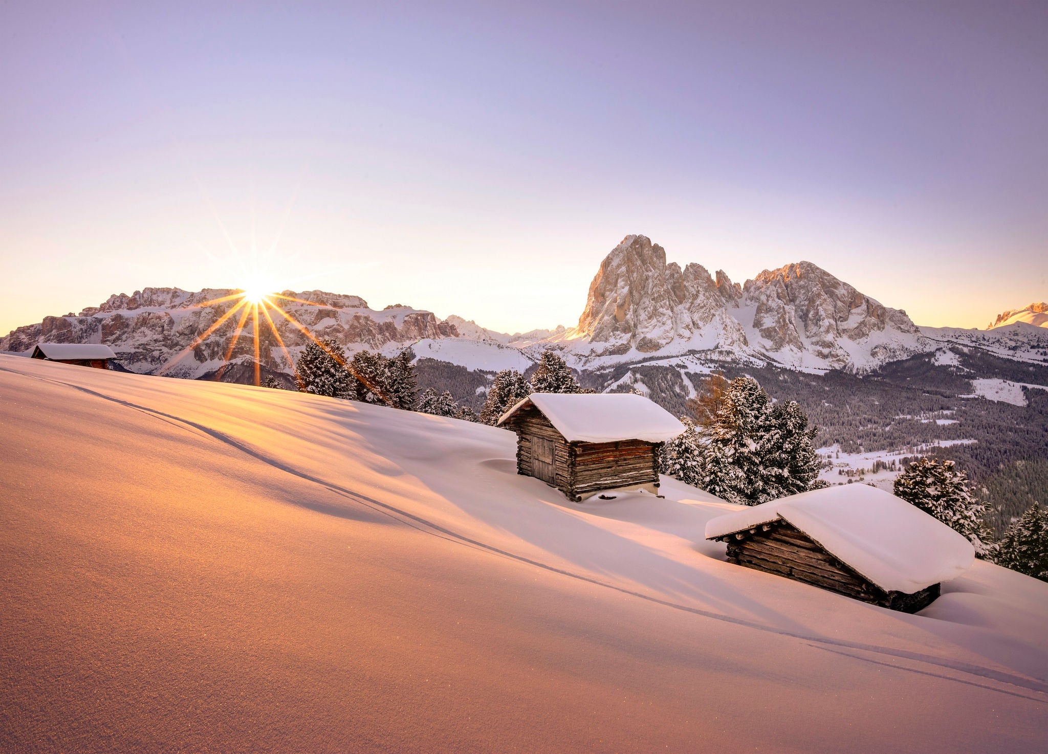 Zwei urige Almhütten mit schneebedecktem Dach werden von der aufgehenden Sonne beleuchtet, dahinter erhebt sich das imposante Dolomitenmassiv der Langkofelgruppe