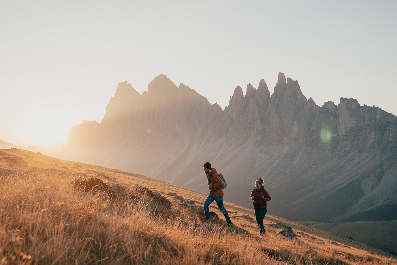 Two people hike in front of the Geisler mountains in Villnösstal with the sun shining from behind them