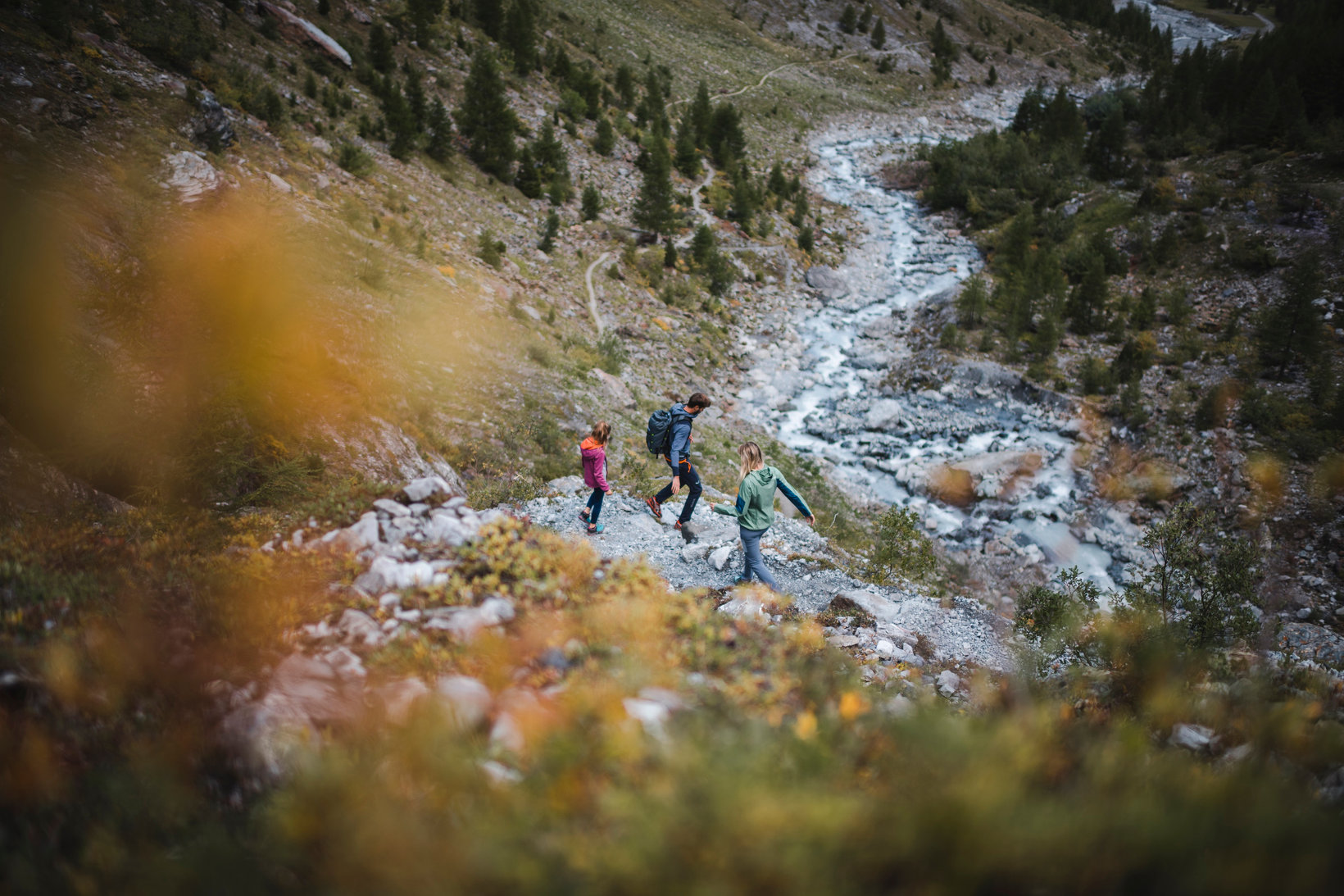 A family of three hikes on a rocky path towards a mountain river in the Stilfserjoch National Park