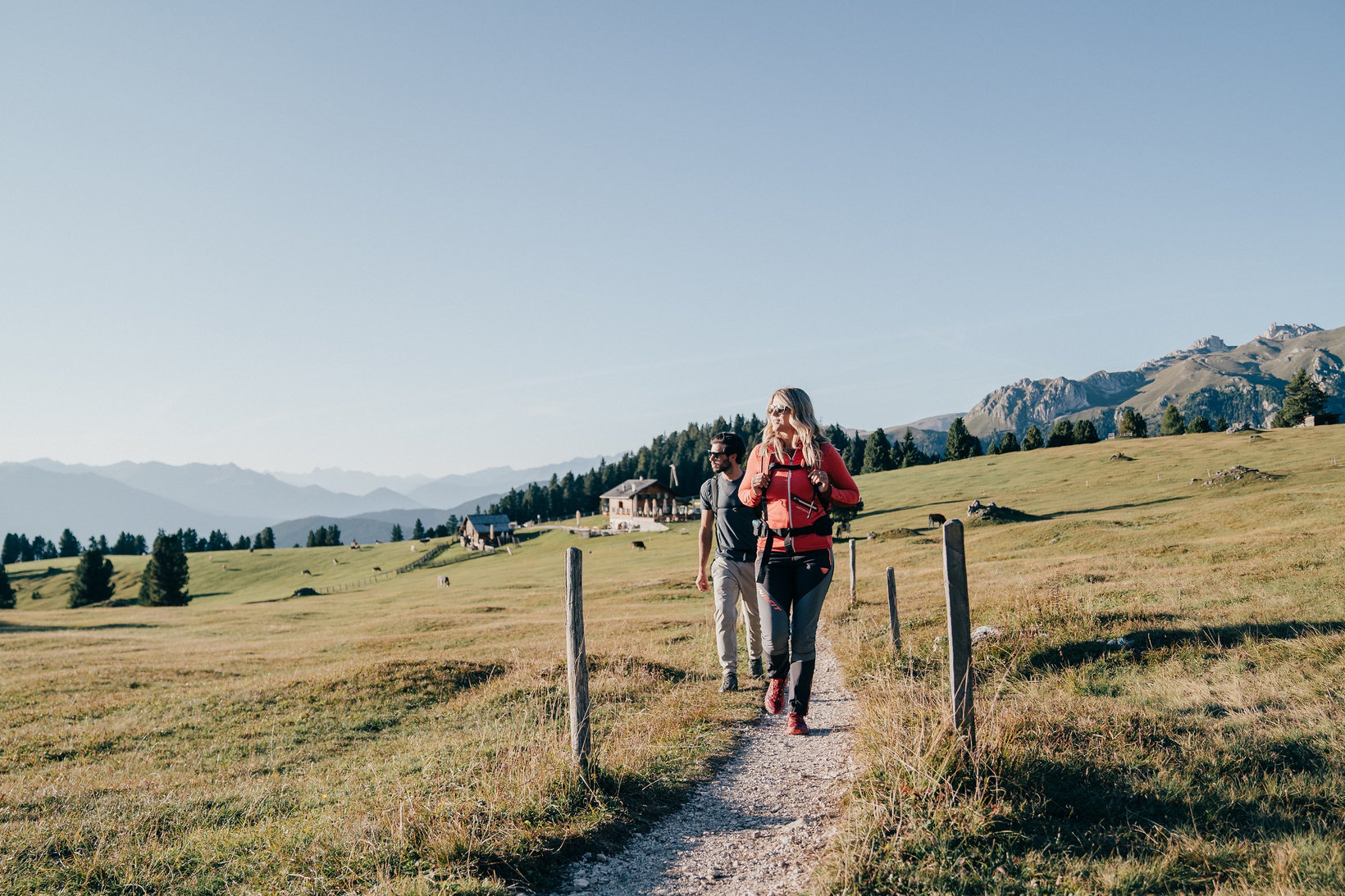 Due persone camminano lungo prati verdi in Val di Funes durante una giornata di sole