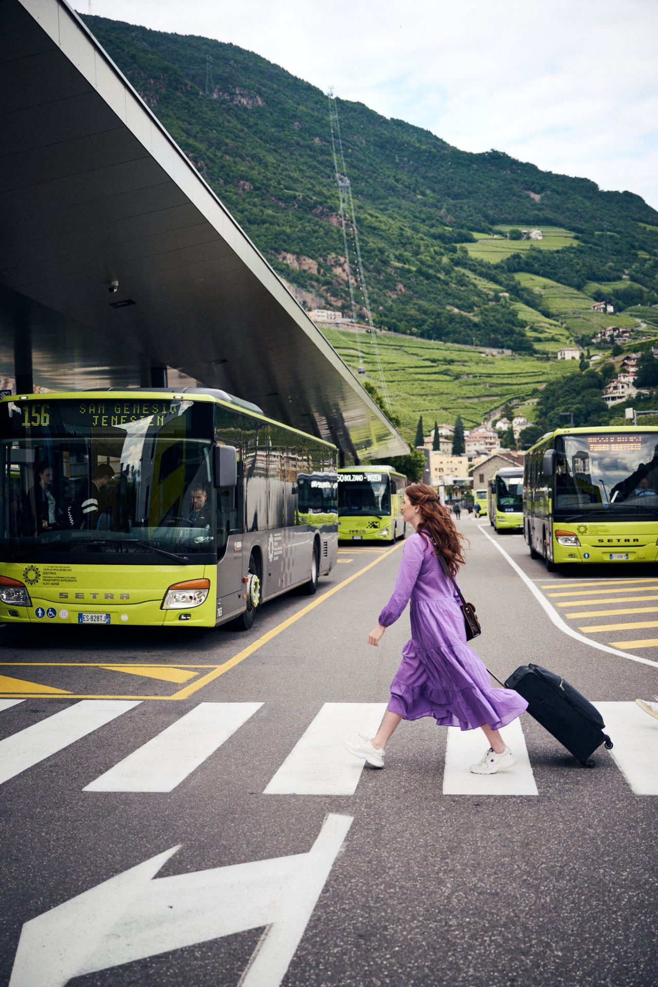 Eine junge Frau zieht ihren kleinen Koffer über einen Zebrastreifen am Busbahnhof in Bozen, mit Blick auf grüne Weinberge.